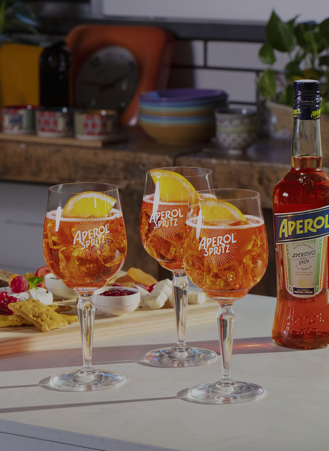 Three Aperol Spritz cocktails garnished with orange slices are placed on a kitchen counter next to a full bottle of Aperol and a wooden platter filled with colorful appetizers, creating a vibrant aperitivo setting.